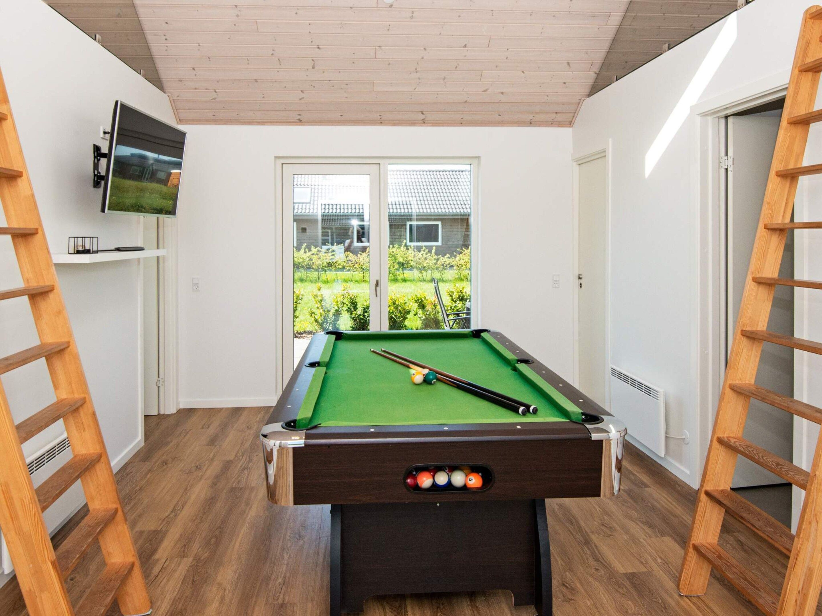 A pool table with a green felt surface, surrounded by wooden flooring, with a television mounted on the wall and a view of a garden through a glass door.