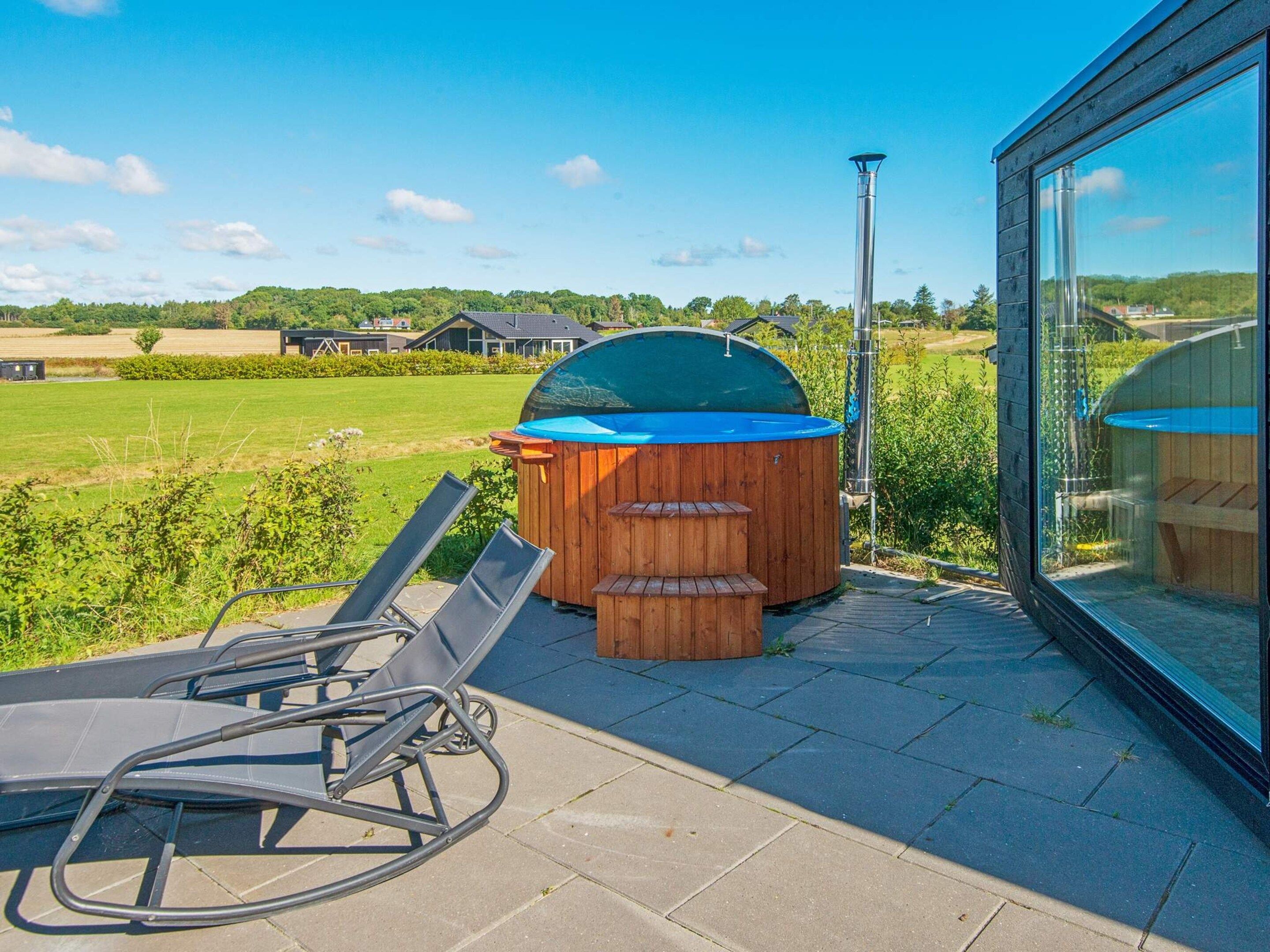 A modern outdoor patio with a hot tub, lounge chairs, and a view of a green landscape.