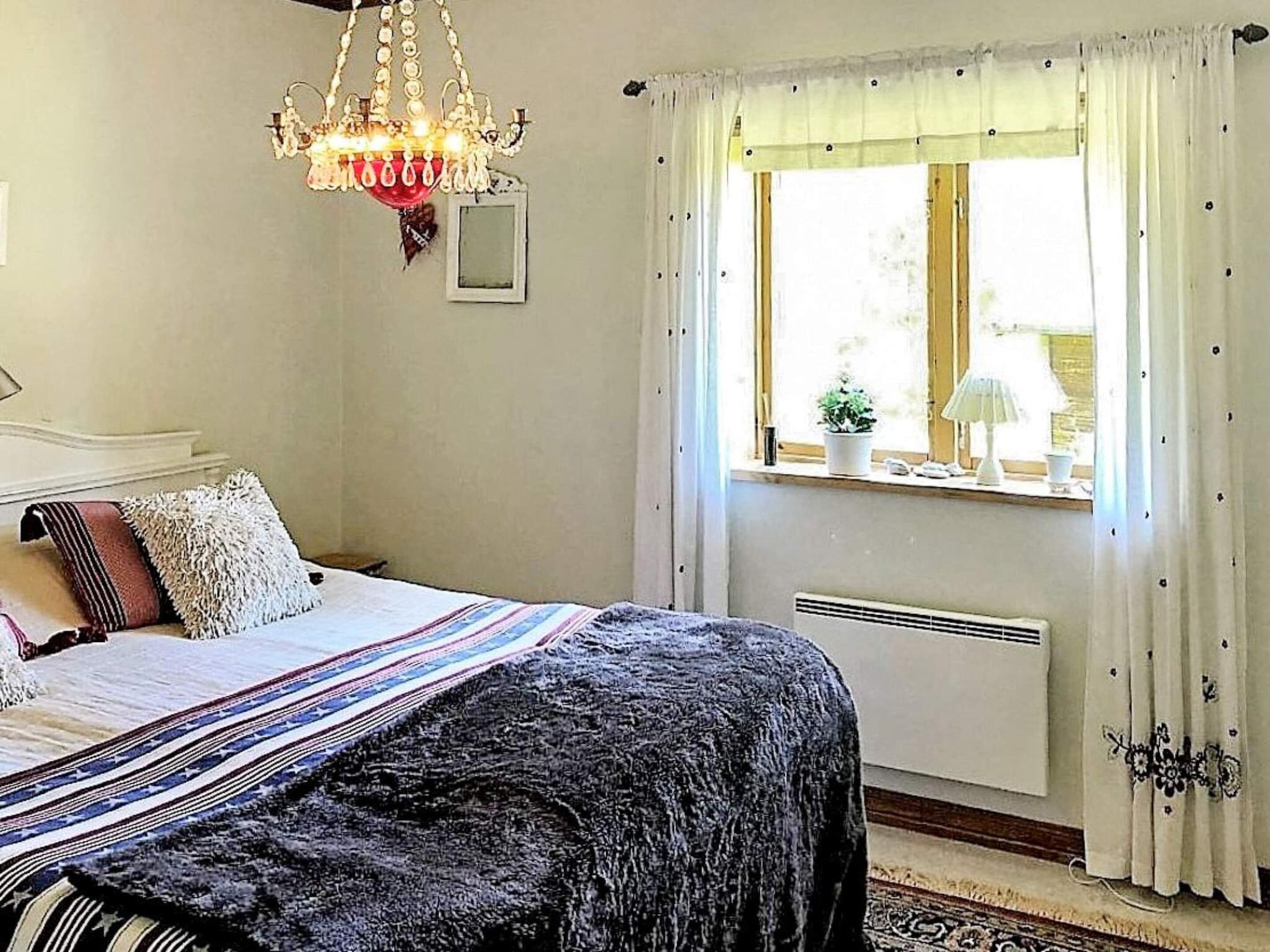 A bedroom with a bed, a chandelier, a window with curtains, a radiator, and a small table with a lamp.