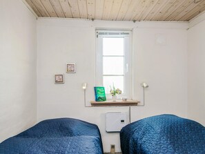 A room with two beds, a window, a small shelf with a book and a vase, and a wall-mounted heater.
