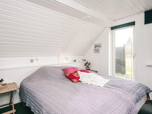Een slaapkamer met een schuine plafond, een bed met een geribbelde deken, een klein tafeltje met een mand, en een raam met uitzicht op buiten.