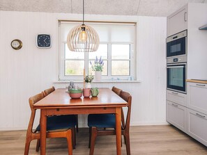 A dining area with a wooden table, chairs, a hanging light fixture, a window with a view, and a kitchen with appliances.