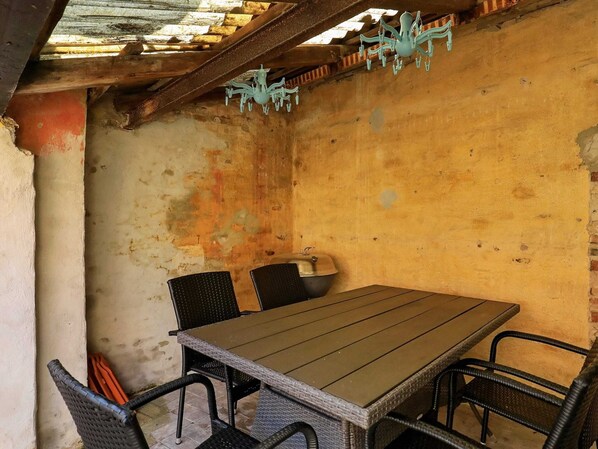 Een rustieke eethoek buiten met een houten tafel en rieten stoelen, een metalen pot op de tafel en een decoratieve hanglamp.