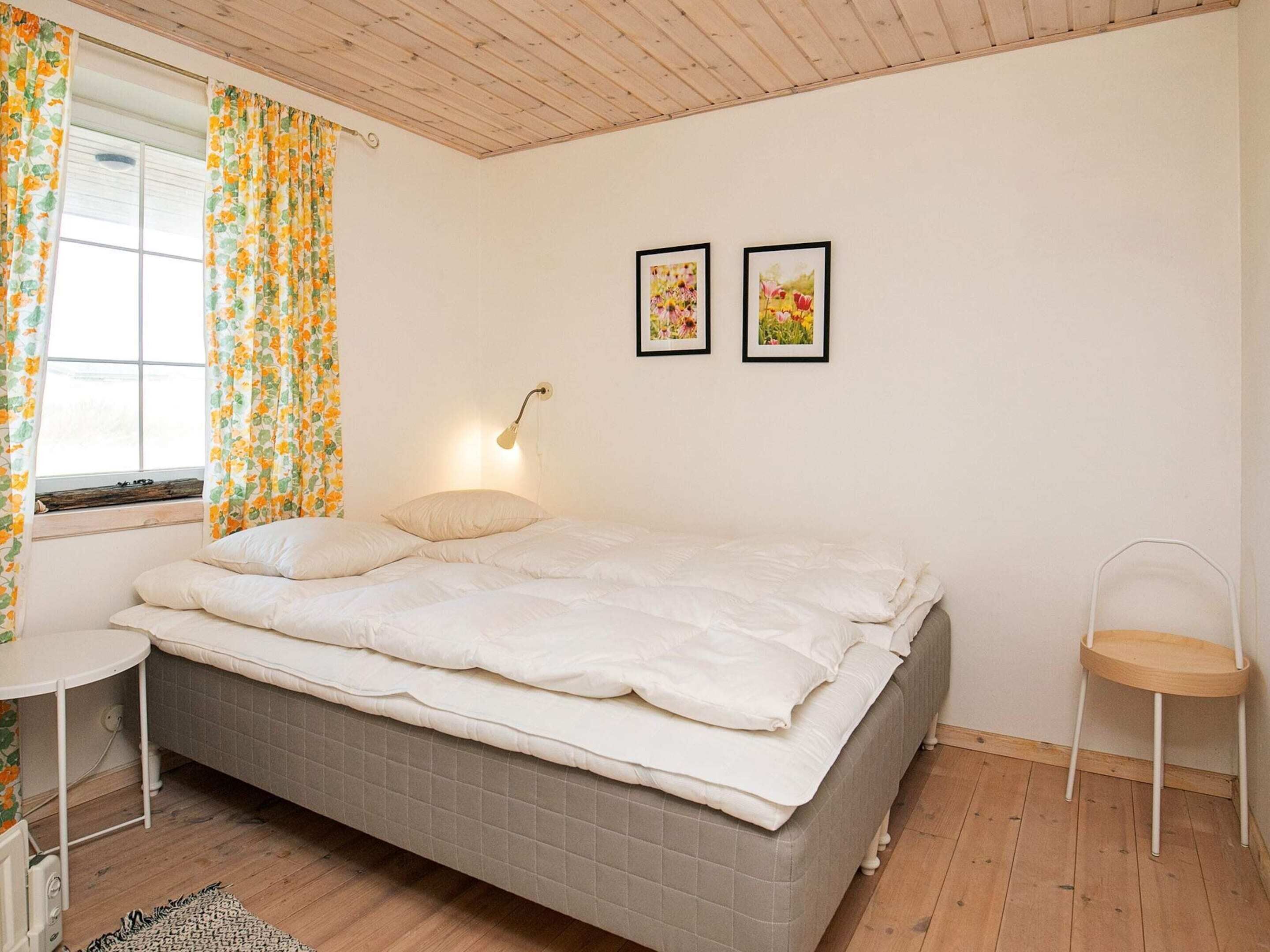 A bedroom with a bed, a bedside table, a chair, a window with curtains, and two framed pictures on the wall.
