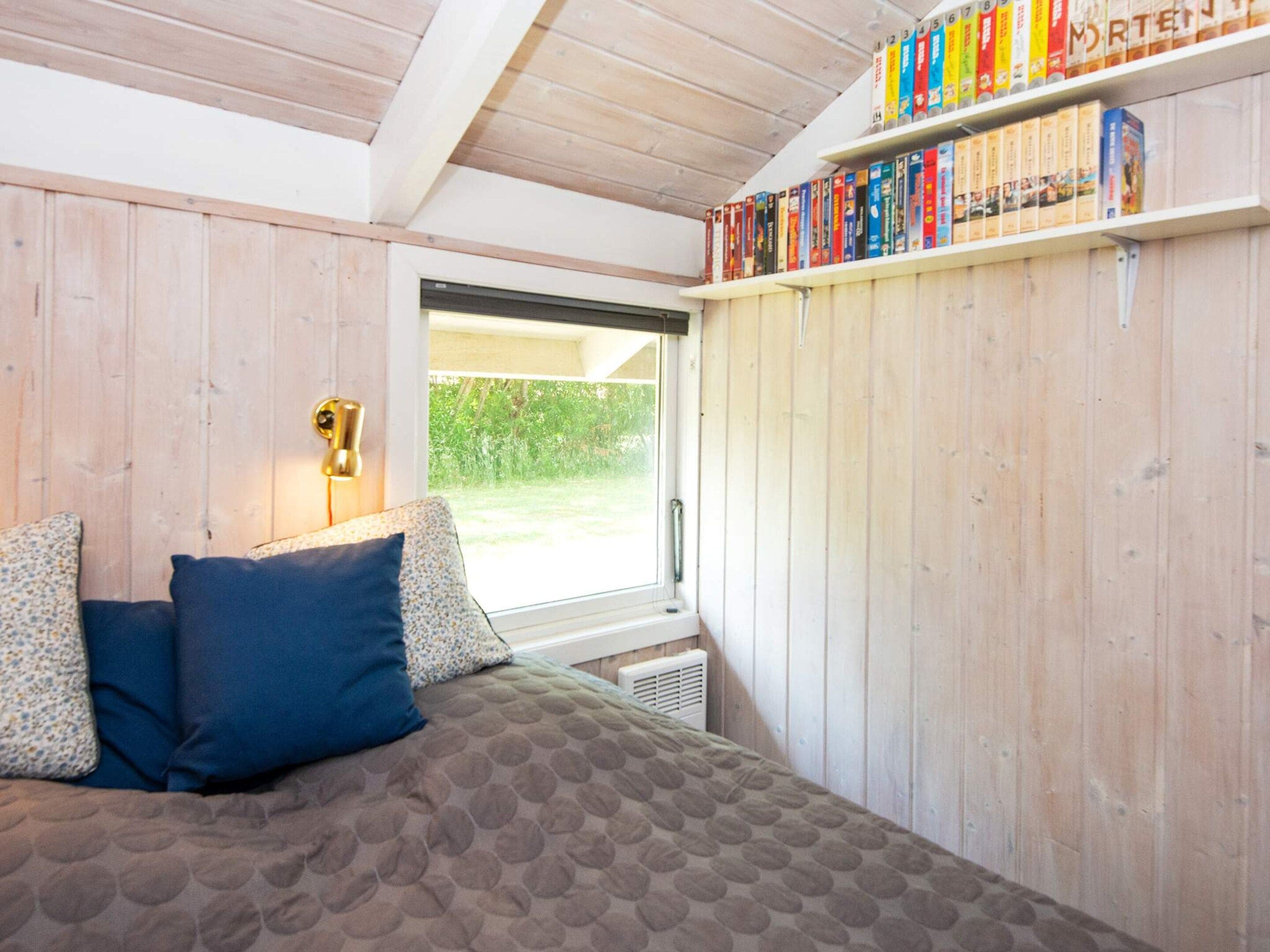 Een slaapkamer met een bed, kussens, een raam en een boekenplank vol boeken.