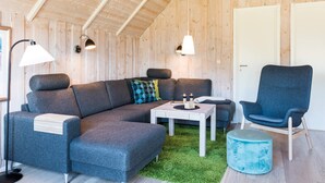 Ein modernes, gemütliches Wohnzimmer mit einer Eckcouch, einem runden Hocker und einem blauen Sessel. Auf einem kleinen Tisch stehen Kerzen und ein Buch. Das Zimmer zeichnet sich durch Holzwände und eine geneigte Decke aus.