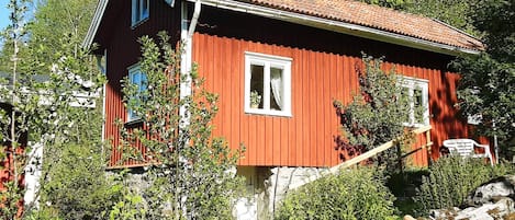 A red wooden house with a sloped roof, surrounded by greenery and rocks.