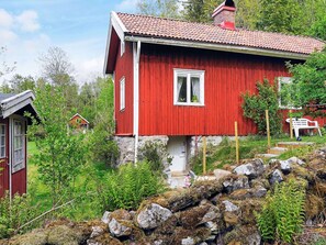 A red wooden house with a stone foundation, surrounded by greenery and rocks.