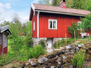 Une maison en bois rouge, avec des fondations en pierre, entourée de verdure et de rochers.