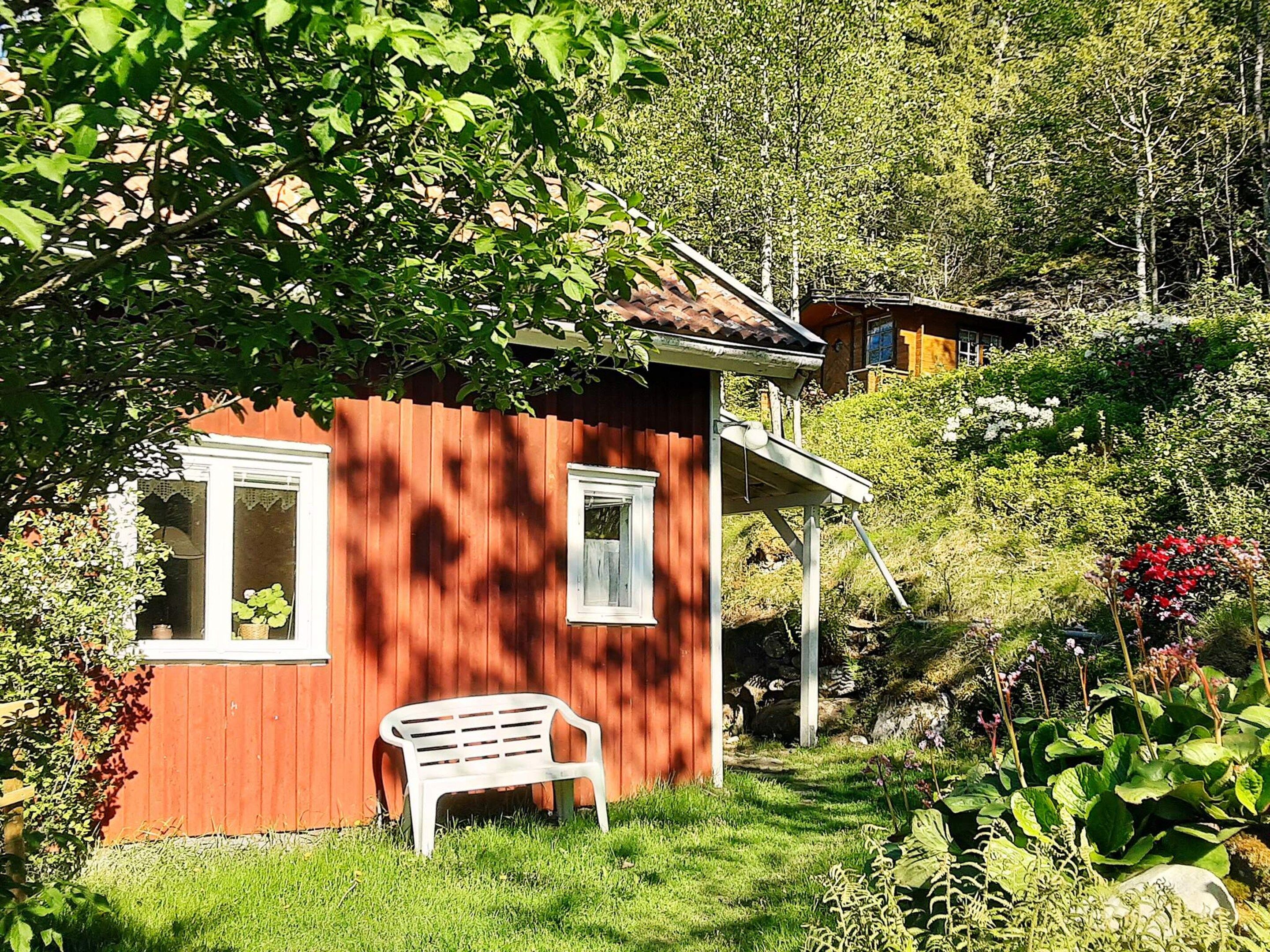 Ett rött trähus med ett vitt fönster, en vit bänk och frodig grönska i bakgrunden.