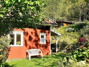 Une maison en bois rouge avec une fenêtre blanche, un banc blanc et une végétation luxuriante en arrière-plan.