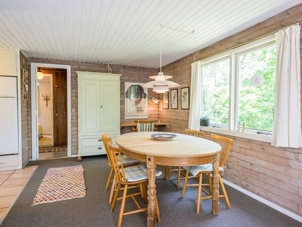 A dining area with a round wooden table, six chairs, a patterned rug, and a window with curtains. - 4 Person Holiday Home in Norre Nebel (Norre Nebel)