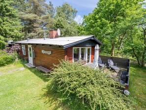 A small wooden cabin with a nameplate, a patio with outdoor furniture, and a surrounding lush green landscape. - 4 Person Holiday Home in Norre Nebel (Norre Nebel)