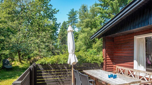 An outdoor patio with a wooden table and chairs, a white umbrella, and a wooden cabin in the background. - 4 Person Holiday Home in Norre Nebel (Norre Nebel)