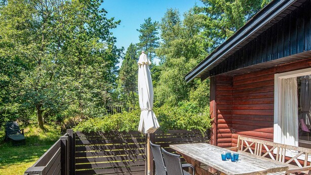 An outdoor patio with a wooden table and chairs, a covered umbrella, and a view of a forested area. - 4 Person Holiday Home in Norre Nebel (Norre Nebel)