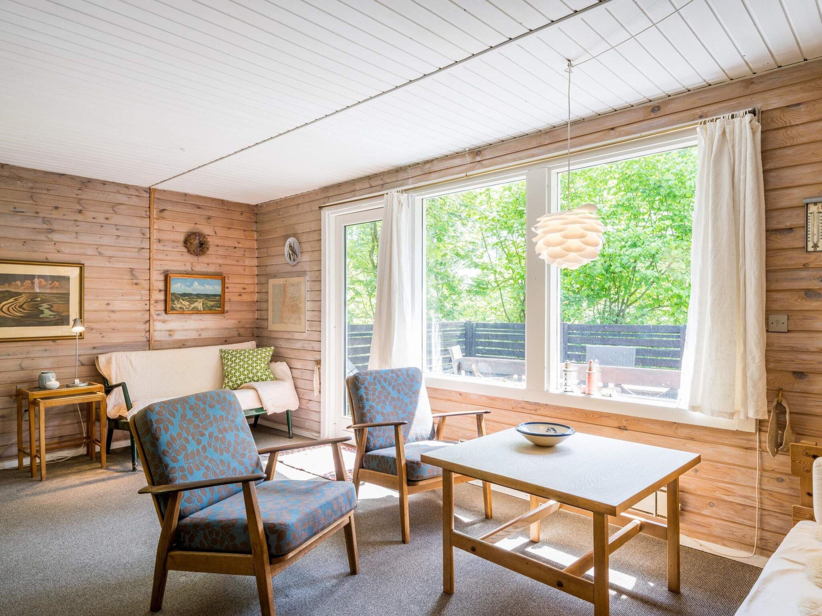 A cozy room with a wooden interior, a sofa, two chairs, a table, and a large window with a view of greenery.
