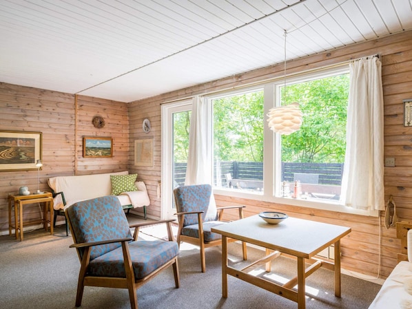 Une pièce chaleureuse avec un décor intérieur en bois, un canapé, deux fauteuils, une table et une grande fenêtre donnant sur un paysage verdoyant.
