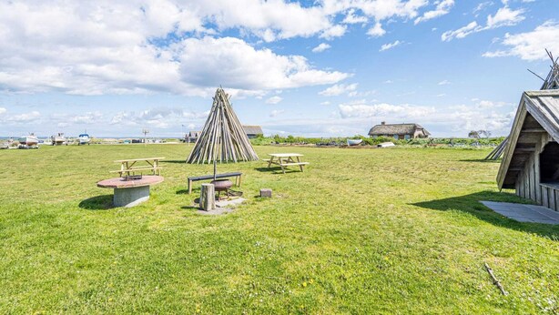A grassy field with a wooden structure, picnic tables, and a thatched-roof building. - 4 Person Holiday Home in Hvide Sande (Hvide Sande)