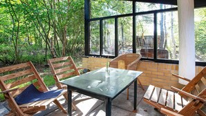 An outdoor patio with a glass table and wooden chairs, surrounded by a brick wall and large windows overlooking a forested area. - 8 Person Holiday Home in Vaeggerlose (Vaeggerlose)