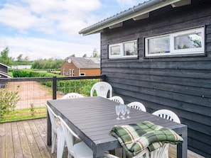 Eine Holzterrasse mit Tisch und Stühlen, ein Haus mit dunkler Fassade und weißer Verzierung sowie ein Blick auf eine grüne Landschaft und ein entferntes Gebäude.