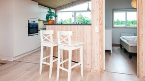 Una cocina moderna con armarios blancos, horno empotrado y ventana con vistas a zonas verdes. Hay dos sillas blancas y una mesita.