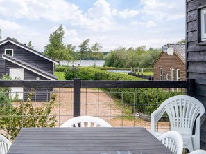 A wooden deck with a table and chairs, overlooking a fenced garden with a lake view. - Holiday Home in Ansager (Ansager)