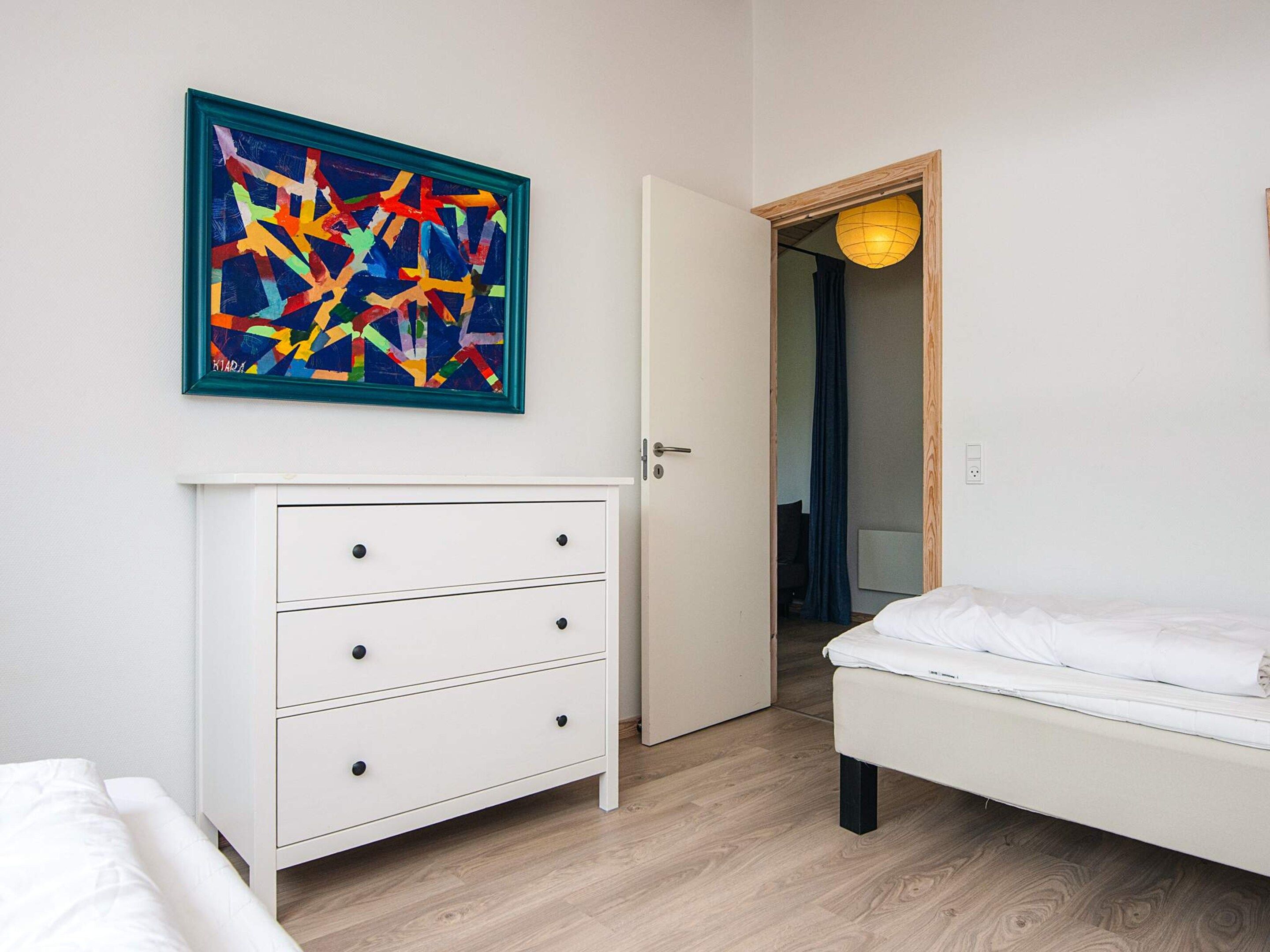 Une chambre avec une commode blanche, un lit et un tableau abstrait aux couleurs vives accroché au mur.