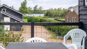 Una terraza de madera con una mesa y sillas, un jardín cercado y vistas a un lago y edificios.