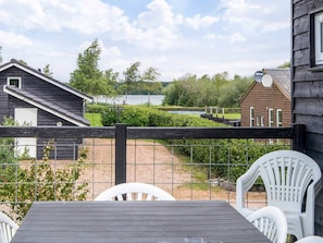Een houten terras met een tafel en stoelen, een omheinde tuin en uitzicht op een meer en gebouwen.