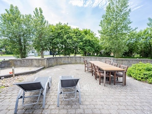 En udendørs patio med stenbelægning, et langt træbord med stole, og metalstole.