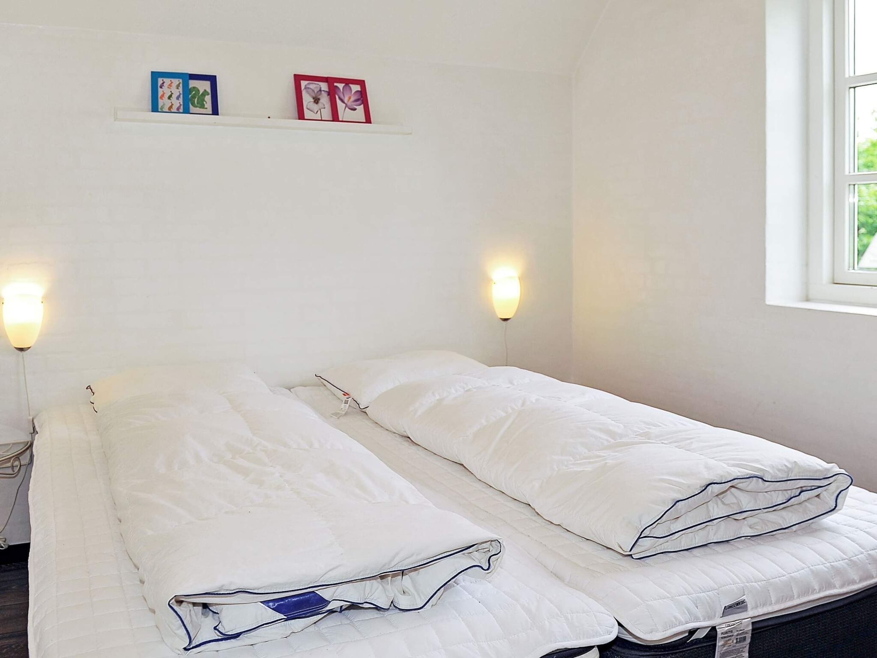 A room with two single beds, each with white bedding and a blue tag. There are wall-mounted light fixtures on either side of the beds. A small shelf above the beds holds two framed pictures.