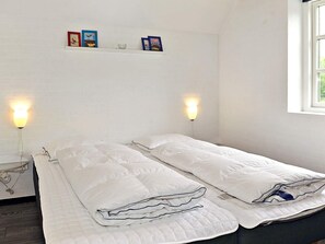 A room with two beds, each with a white quilted mattress and pillows, a small shelf with framed pictures, and wall-mounted lamps.