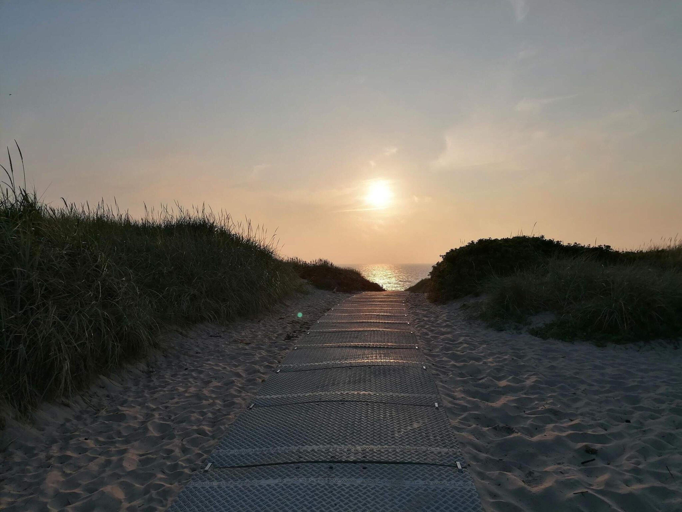 A pathway leading to the sea with a metal grid surface, flanked by grass on both sides.
