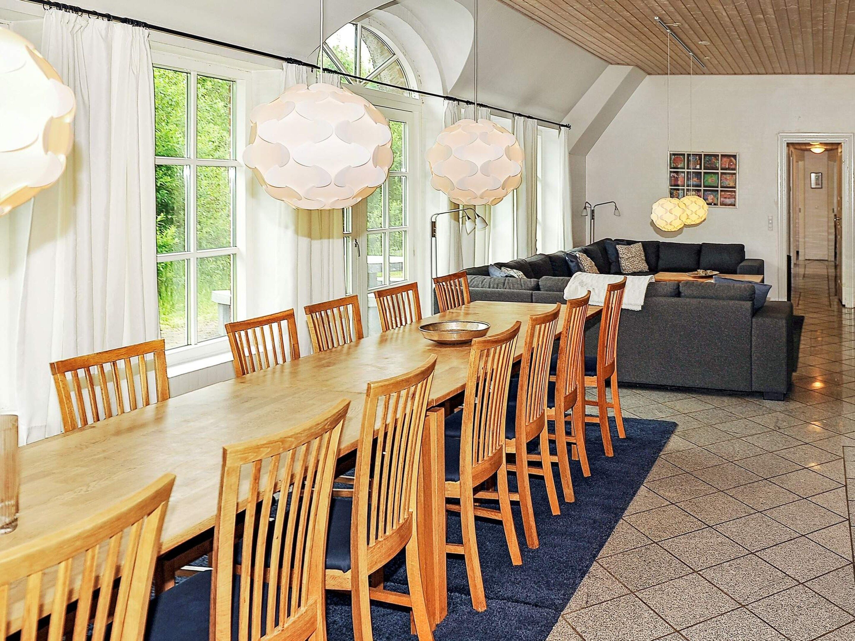 A dining area with a long wooden table and chairs, a sofa, and large windows with curtains.