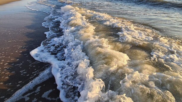 Uma praia com ondas quebrando na costa, céu claro e uma linha de costa distante.