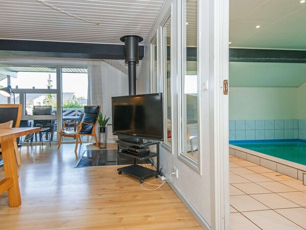 Una sala de estar moderna con televisor de pantalla plana, muebles de madera, un ventanal que da a una piscina y un área exterior visible.