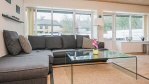 A modern living room with a grey sectional sofa, a glass coffee table, and large windows offering a view of the outside. - 4 Person Holiday Home in Norre Nebel (Norre Nebel)