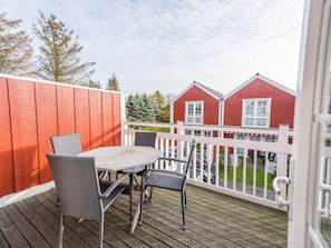 Une terrasse en bois avec une table et des chaises, un bâtiment rouge en arrière-plan et une clôture blanche.