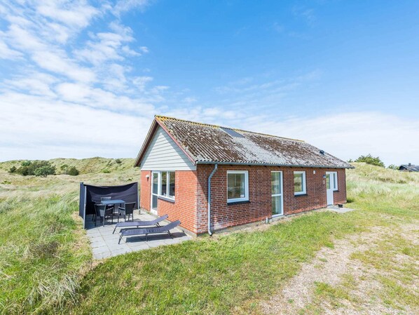 A small brick house with a sloped roof, a covered outdoor seating area, and a gravel path leading to the entrance.