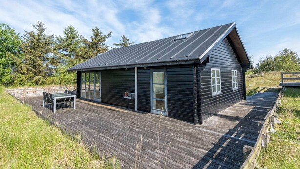 Eine schwarze Holzhütte mit geneigtem Dach, einer Terrasse und einem Tisch mit Stühlen davor.