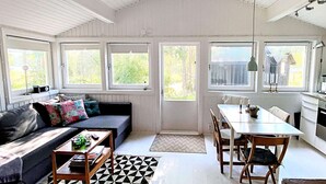 A cozy living room with a sofa, coffee table, and dining area with chairs. There are large windows providing natural light and views of the outdoors.