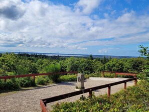 Ein Kiesweg mit Holzzaun und Steinmauer, der zu einer Aussichtsplattform mit Blick auf eine bewaldete Landschaft und ein entferntes Gewässer unter teilbewölktem Himmel führt.