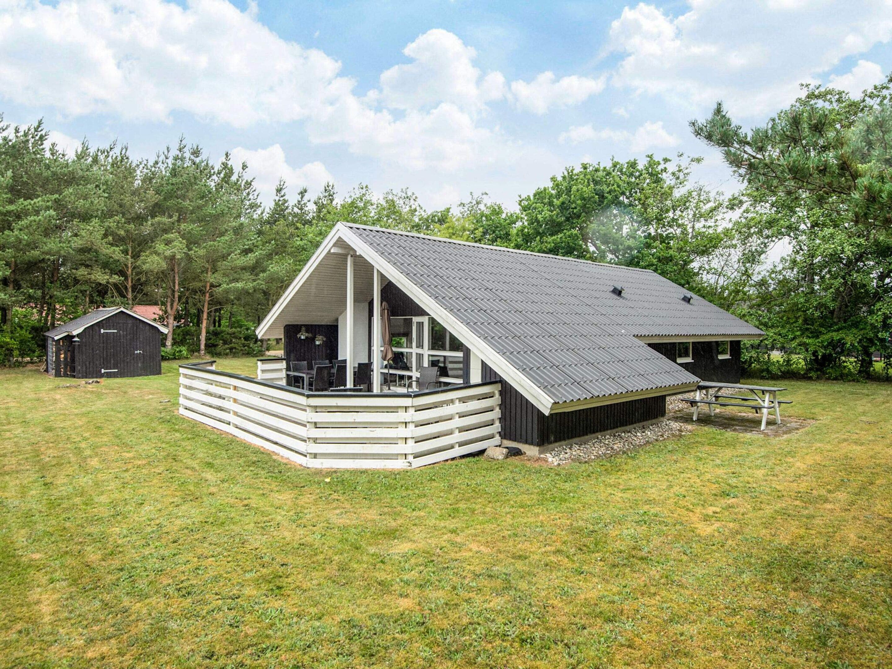 A single-story cabin with a porch, a picnic table, and a small shed in a grassy area.