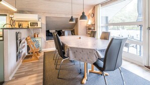 A modern kitchen with wooden cabinetry, a dining area with a large table and chairs, and a view through a large window. - 4 Star Holiday Home in Norre Nebel (Norre Nebel)