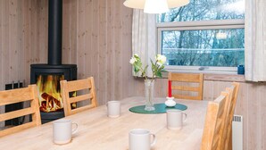 Ett mysigt rum med en vedeldad kamin, ett matbord med stolar, en vas med blommor och ett fönster med utsikt över träd.
