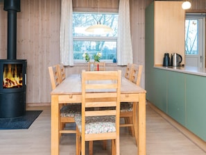 A cozy room with a wood-burning stove, a dining table with chairs, a window with curtains, and a kitchen area with cabinets and appliances.