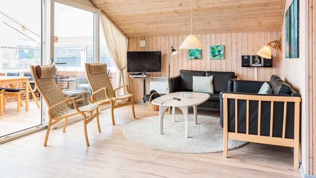 A spacious living area with wooden flooring, a round coffee table, a sofa, and two armchairs. A flat-screen TV is mounted on the wall, and there are two wall-mounted lamps. - 4 Person Holiday Home in Henne (Henne)