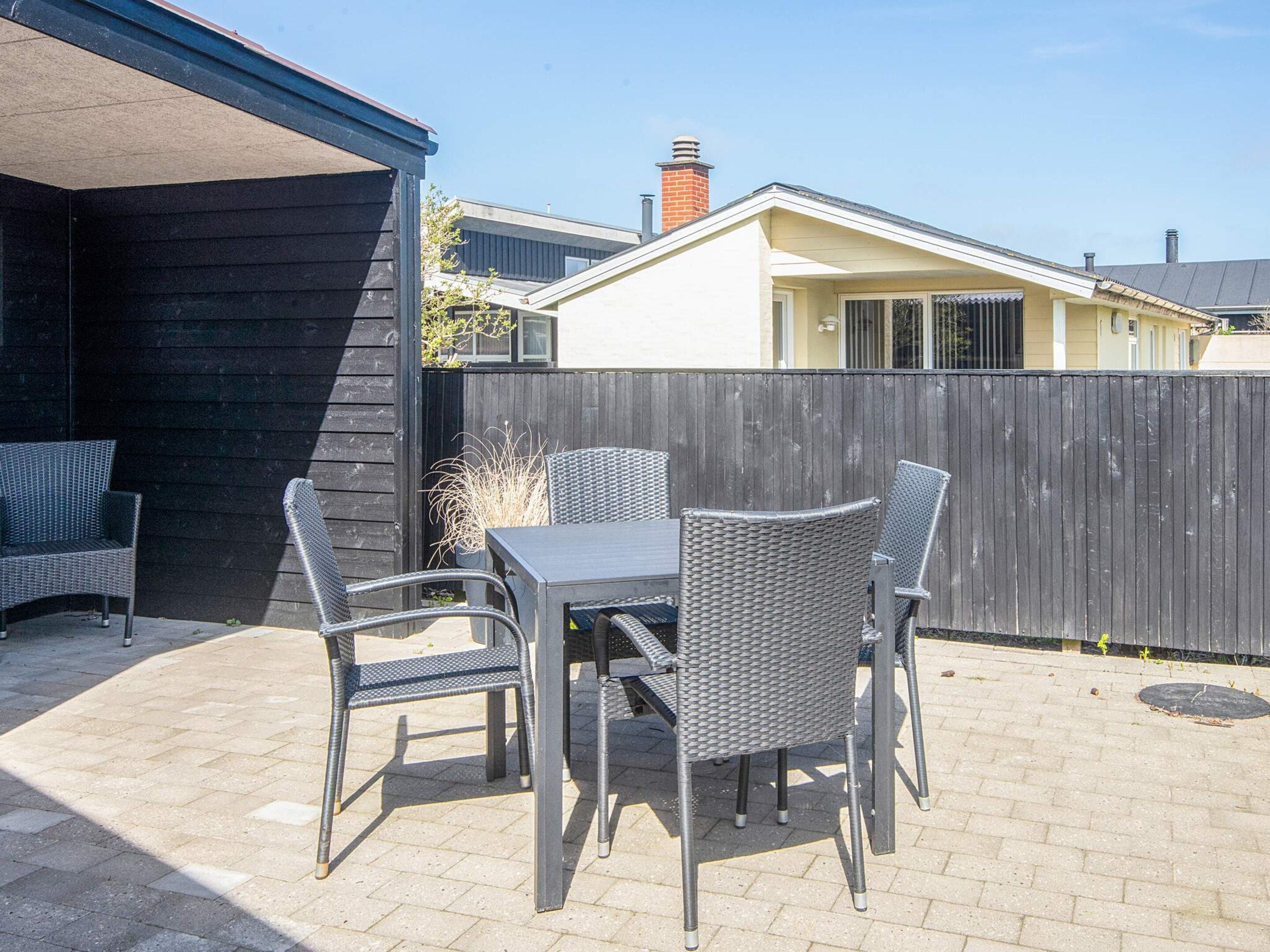 An outdoor patio with a table and four chairs, a wooden fence, and a building in the background.