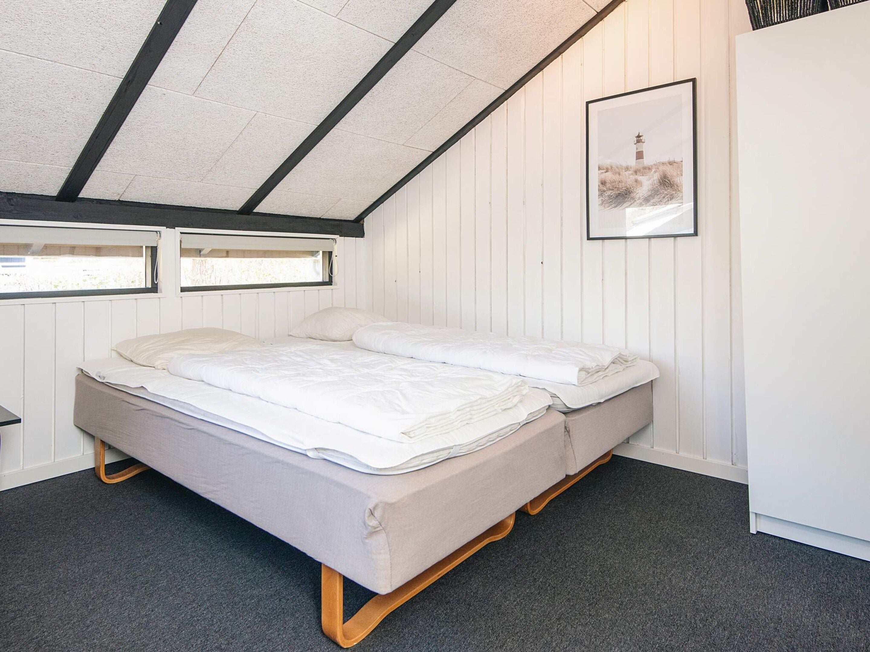 En enkeltseng med træramme og grå madras i et rum med hvide vægge og et skråt loft.