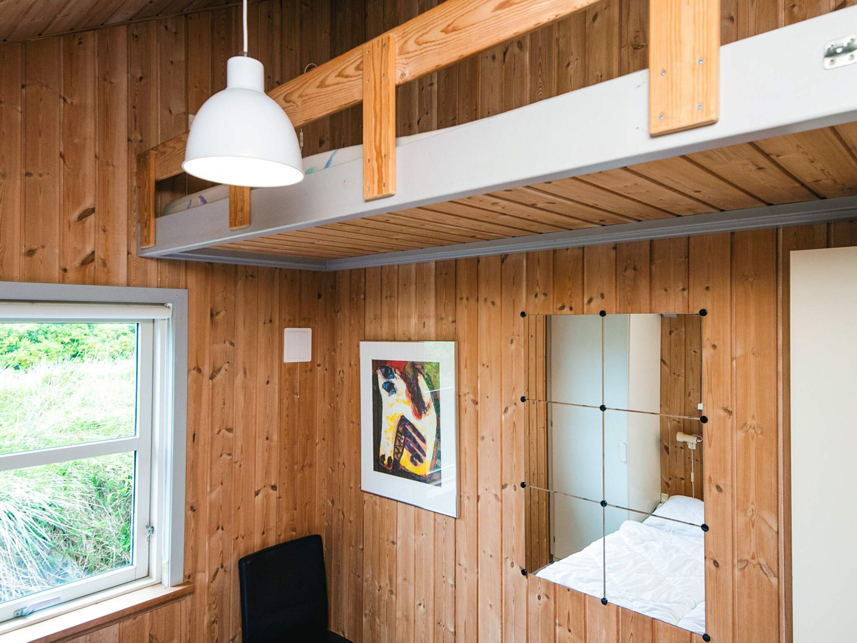 A room with wooden walls, a bed with a white mattress, a black chair, a white pendant light, and a window with a view of greenery.